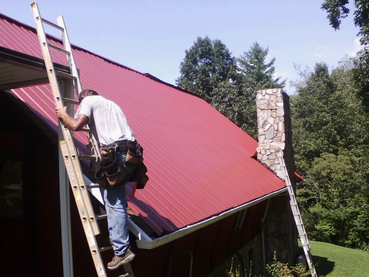 Roofer on a ladder beside a red metal roof and stone chimney — Soffit & Fascia Repair by Roofing Brothers in Peabody